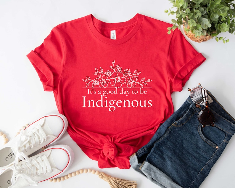 May include: Red t-shirt with a white floral design and the text "It's a good day to be Indigenous".