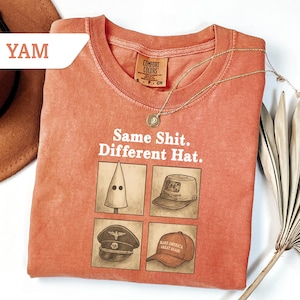 Peut inclure: Un t-shirt corail avec le texte "Same Shit. Different Hat." et quatre illustrations de chapeaux. Un collier en or et un chapeau marron sont &eacute;galement visibles. Le t-shirt est de Comfort Colors.