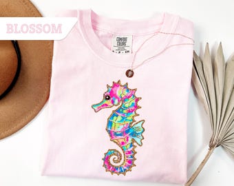 May include: A light pink t-shirt with a colorful seahorse design. The seahorse is decorated with blue, green, pink, and yellow hues, outlined with gold glitter. The word "BLOSSOM" is printed on a white banner. A gold necklace is draped on the shirt.