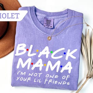May include: A violet-coloured t-shirt with the text "BLACK MAMA" in white, with colourful dots above the letters. The shirt also has the phrase "I'M NOT ONE OF YOUR LIL FRIENDS" in white. A gold necklace is draped over the shirt.