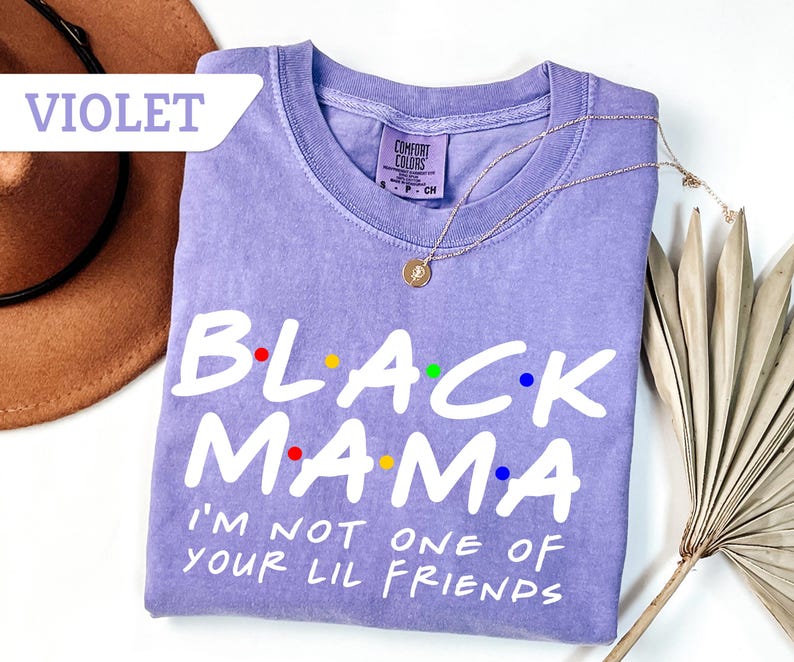 May include: A violet-coloured t-shirt with the text "BLACK MAMA" in white, with colourful dots above the letters. The shirt also has the phrase "I'M NOT ONE OF YOUR LIL FRIENDS" in white. A gold necklace is draped over the shirt.