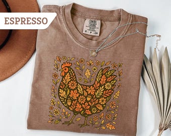 May include: Espresso-coloured t-shirt with a floral rooster design in orange, yellow, and green. The shirt has a crew neck and short sleeves. A silver necklace with a pendant rests on the shirt. The word "ESPRESSO" is in a white banner in the upper left corner.