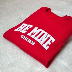 May include: A folded red sweatshirt with the words "BE MINE" in large white block letters. Below the text is the word "VALENTINE" in a white rectangular box. The sweatshirt is on a white, fluffy surface.