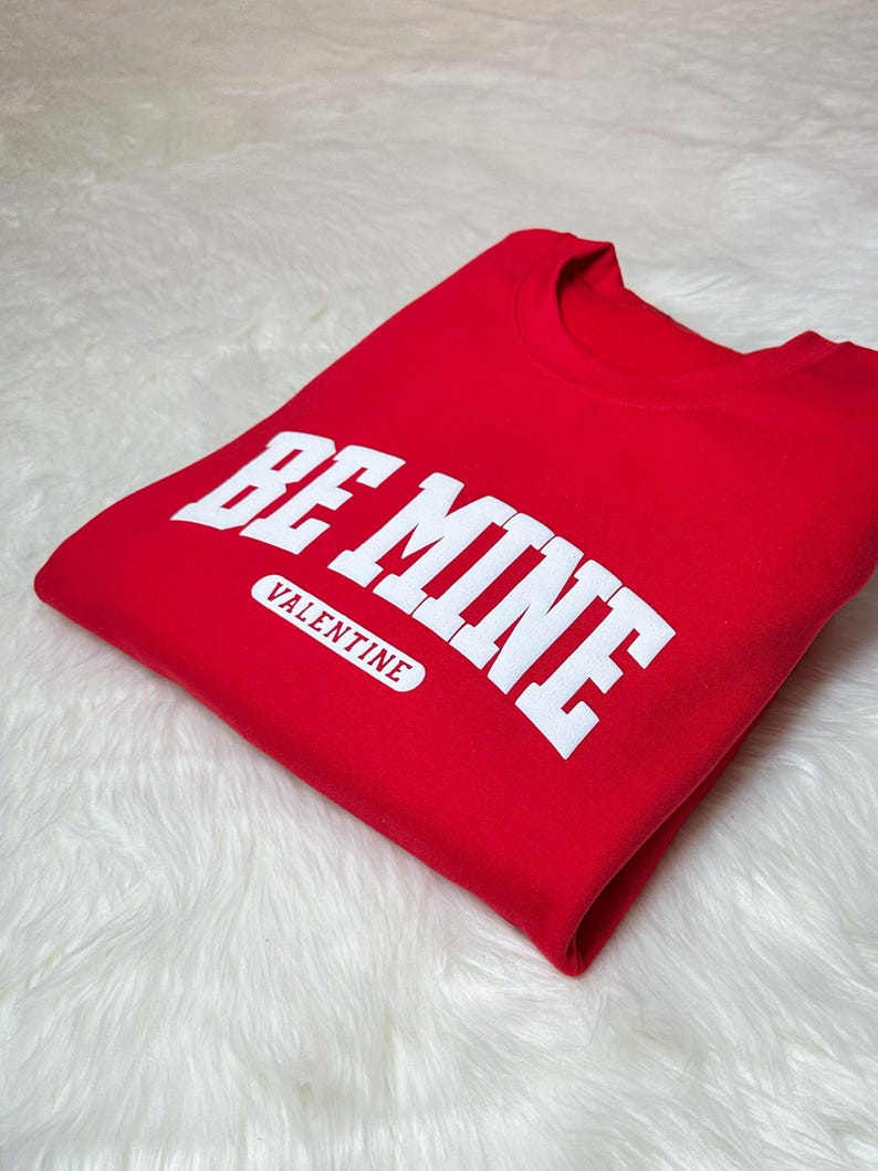 May include: A folded red sweatshirt with the words "BE MINE" in large white block letters. Below the text is the word "VALENTINE" in a white rectangular box. The sweatshirt is on a white, fluffy surface.