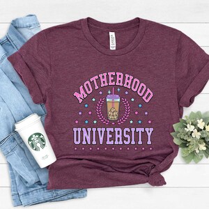 May include: A maroon t-shirt with the text "MOTHERHOOD UNIVERSITY" in pink and purple lettering. A cartoon iced coffee cup with a pink straw and ice cubes is in the center of the text. The text is surrounded by pink and blue stars.
