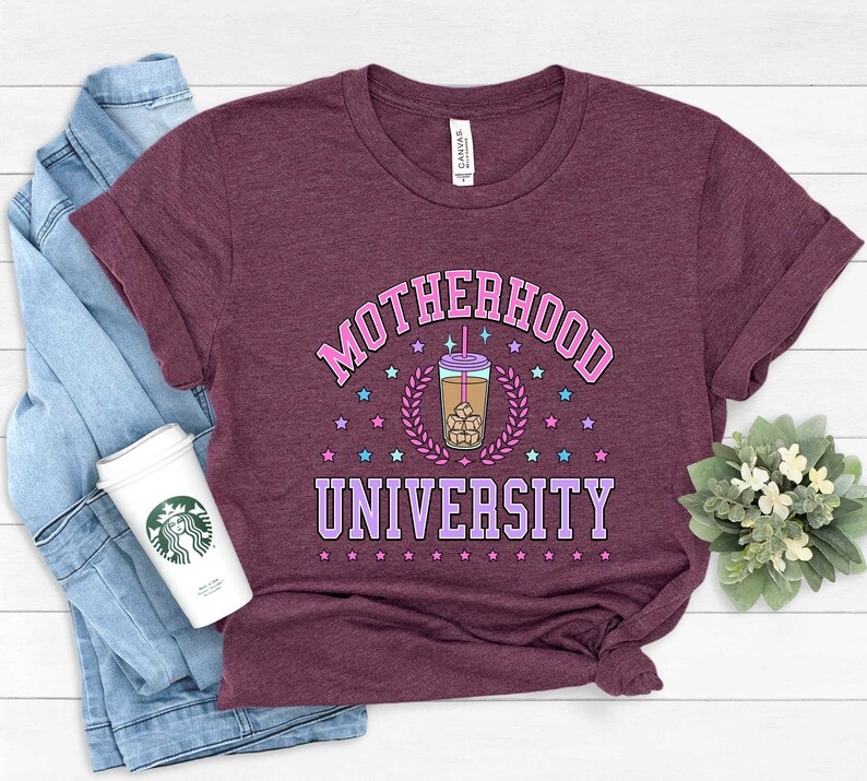 May include: A maroon t-shirt with the text "MOTHERHOOD UNIVERSITY" in pink and purple lettering. A cartoon iced coffee cup with a pink straw and ice cubes is in the center of the text. The text is surrounded by pink and blue stars.