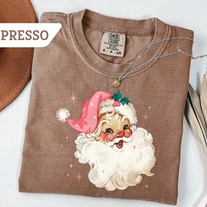 May include: Espresso-colored t-shirt featuring a vintage Santa Claus graphic. The Santa Claus illustration has a pink hat, white beard, and rosy cheeks. A silver necklace with a pendant is draped over the shirt. The shirt is a Comfort Colors brand.