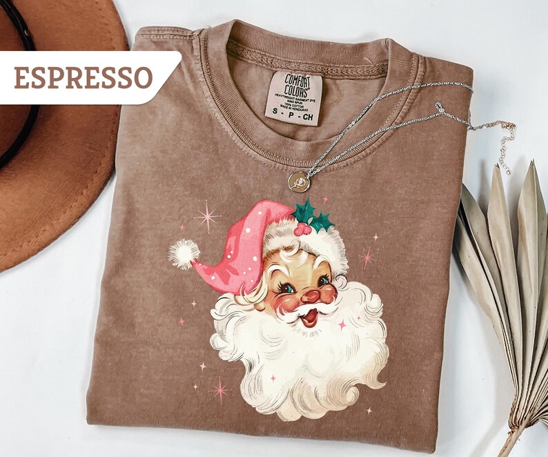 May include: Espresso-colored t-shirt featuring a vintage Santa Claus graphic. The Santa Claus illustration has a pink hat, white beard, and rosy cheeks. A silver necklace with a pendant is draped over the shirt. The shirt is a Comfort Colors brand.