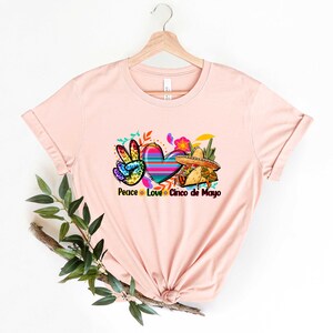 Puede incluir: Una camiseta rosa con un gr&aacute;fico de un signo de paz, un coraz&oacute;n y un sombrero. El texto "Peace Love Cinco de Mayo" est&aacute; impreso debajo del gr&aacute;fico.