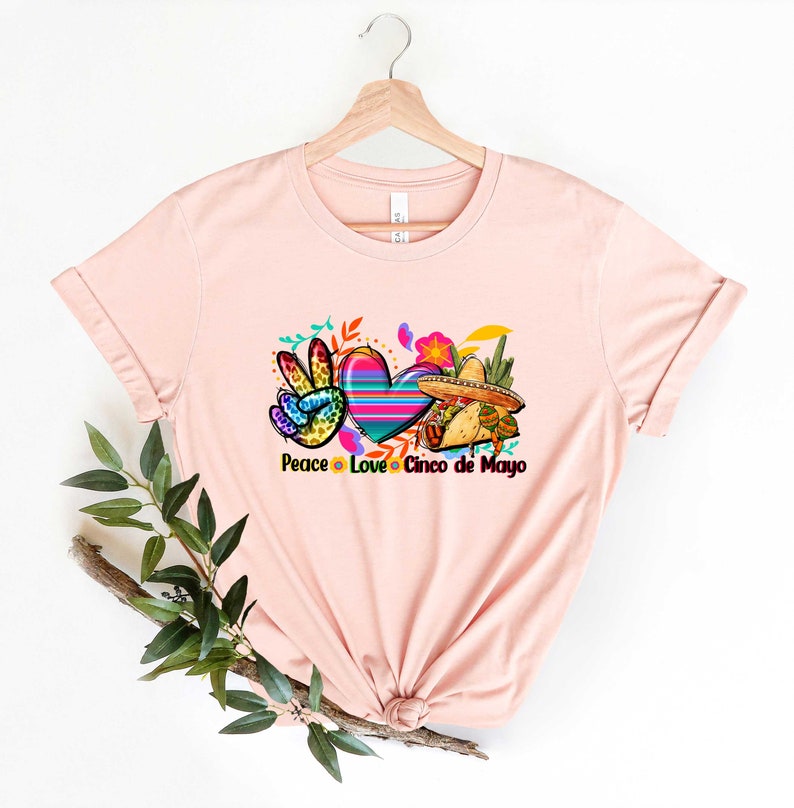 Puede incluir: Una camiseta rosa con un gr&aacute;fico de un signo de paz, un coraz&oacute;n y un sombrero. El texto "Peace Love Cinco de Mayo" est&aacute; impreso debajo del gr&aacute;fico.