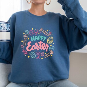 May include: A blue sweatshirt with a white and pink graphic design that says "Happy Easter" surrounded by colorful Easter eggs, carrots, and flowers.
