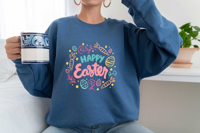 May include: A blue sweatshirt with a white and pink graphic design that says "Happy Easter" surrounded by colorful Easter eggs, carrots, and flowers.