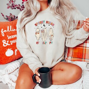 May include: A beige sweatshirt with a graphic of three ghosts holding various items. The ghosts are labeled "OT", "SLP", and "PT". The text "Spooky Squad" is above the ghosts.
