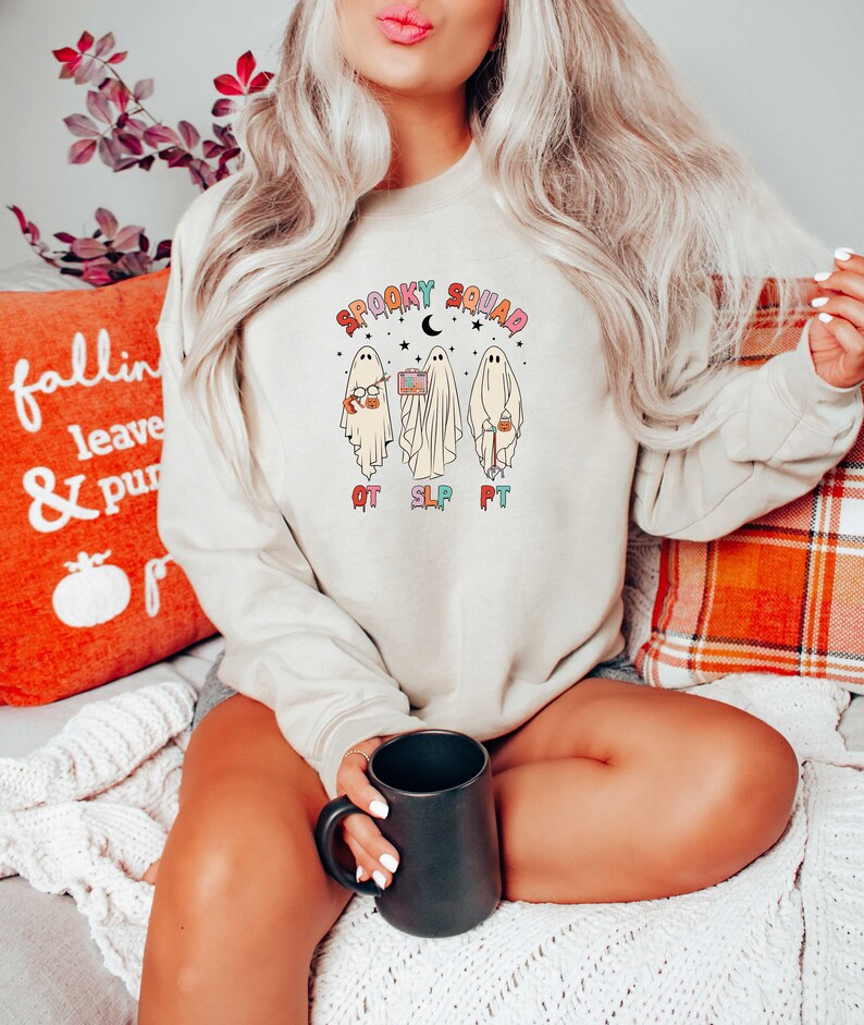 May include: A beige sweatshirt with a graphic of three ghosts holding various items. The ghosts are labeled "OT", "SLP", and "PT". The text "Spooky Squad" is above the ghosts.
