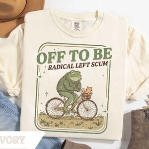 May include: Ivory-colored t-shirt with a green and brown graphic. The graphic features a frog riding a bicycle with a cat in the basket, and the text "OFF TO BE RADICAL LEFT SCUM". The shirt is made of a soft material.