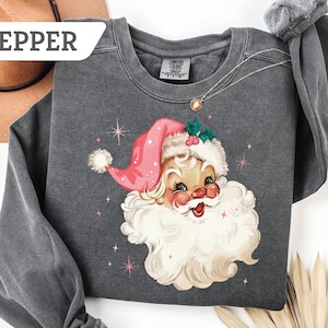 May include: A dark gray sweatshirt featuring a vintage illustration of Santa Claus wearing a pink hat and a white beard. The design includes pink stars and the word "PEPPER" in a white banner. A gold necklace is visible.
