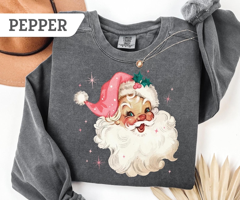 May include: A dark gray sweatshirt featuring a vintage illustration of Santa Claus wearing a pink hat and a white beard. The design includes pink stars and the word "PEPPER" in a white banner. A gold necklace is visible.