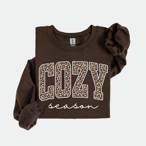 Pu&ograve; includere: Una felpa marrone scuro con la scritta "COZY season". La parola "COZY" &egrave; in grandi lettere con un motivo leopardato, e "season" &egrave; scritto in corsivo.