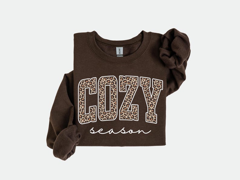 Pu&ograve; includere: Una felpa marrone scuro con la scritta "COZY season". La parola "COZY" &egrave; in grandi lettere con un motivo leopardato, e "season" &egrave; scritto in corsivo.
