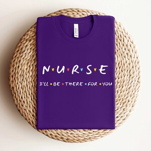 Puede incluir: Camiseta morada con el texto "NURSE" con corazones de colores y el texto "I'll be there for you" en un estilo de fuente similar.