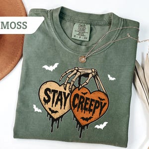 May include: Moss green t-shirt with a skeleton hand holding two heart-shaped pumpkins that read "Stay Creepy". The shirt has a crew neck and short sleeves. White bat graphics are scattered around the design. A gold necklace is draped across the shirt.