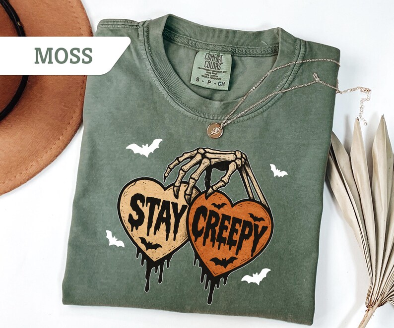 May include: Moss green t-shirt with a skeleton hand holding two heart-shaped pumpkins that read "Stay Creepy". The shirt has a crew neck and short sleeves. White bat graphics are scattered around the design. A gold necklace is draped across the shirt.