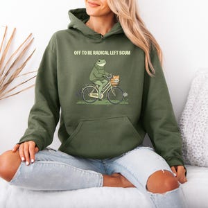 May include: Olive green hoodie with a graphic of a frog riding a bicycle with a cat in the basket. The text "OFF TO BE RADICAL LEFT SCUM" is printed above the graphic. The hoodie has a kangaroo pocket and a drawstring hood.