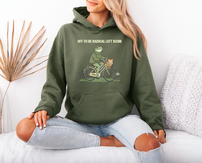 May include: Olive green hoodie with a graphic of a frog riding a bicycle with a cat in the basket. The text "OFF TO BE RADICAL LEFT SCUM" is printed above the graphic. The hoodie has a kangaroo pocket and a drawstring hood.