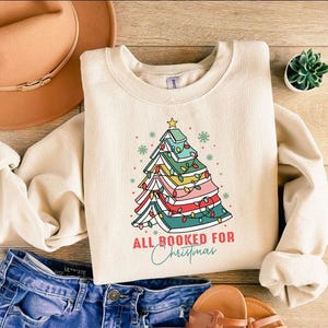 May include: Cream-colored sweatshirt with a Christmas tree design made of books, fairy lights, and a star. The text "ALL BOOKED FOR Christmas" is below the tree. The sweatshirt is paired with blue jeans and brown sandals.