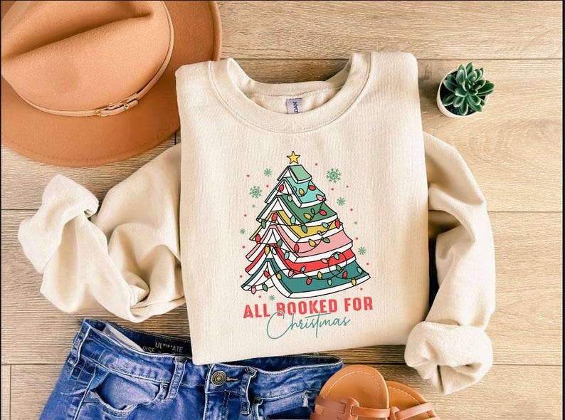 May include: Cream-colored sweatshirt with a Christmas tree design made of books, fairy lights, and a star. The text "ALL BOOKED FOR Christmas" is below the tree. The sweatshirt is paired with blue jeans and brown sandals.