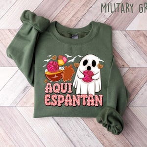 May include: A green sweatshirt featuring a ghost holding a pink pastry and the text "Aqui Espantan" in pink lettering.