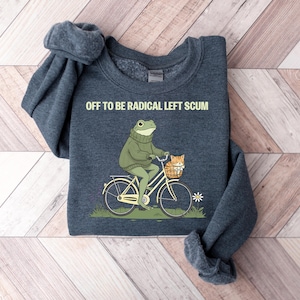 May include: Dark blue sweatshirt with the text "OFF TO BE RADICAL LEFT SCUM." The graphic shows a frog riding a bicycle with a cat in the basket. The frog wears a green sweater. The bicycle is cream-colored.