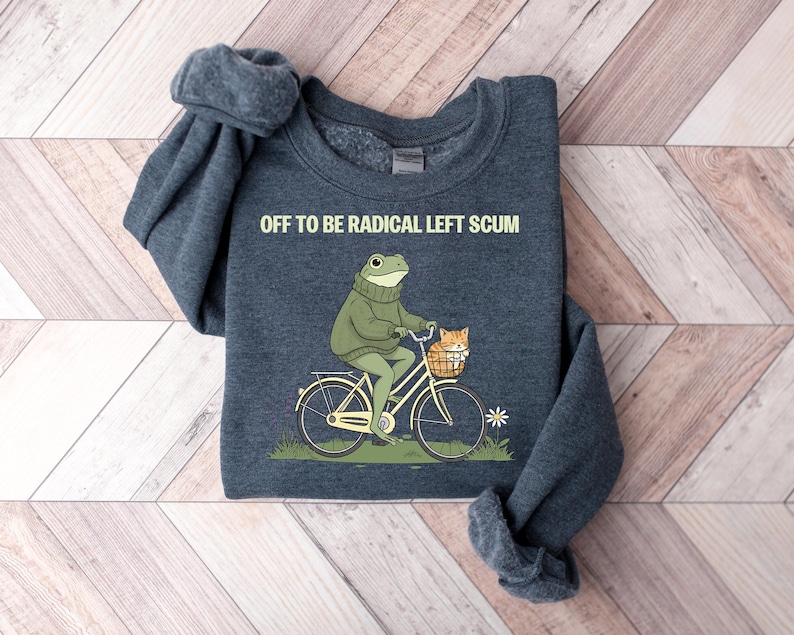 May include: Dark blue sweatshirt with the text "OFF TO BE RADICAL LEFT SCUM." The graphic shows a frog riding a bicycle with a cat in the basket. The frog wears a green sweater. The bicycle is cream-colored.