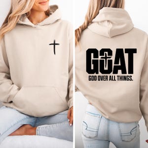 Pode incluir: Um moletom bege com bolso frontal e capuz. Uma cruz preta &eacute; impressa na frente. A parte de tr&aacute;s do moletom apresenta a palavra "GOAT" com uma cruz e o texto "GOD OVER ALL THINGS."