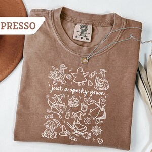 May include: Espresso-colored t-shirt with a white graphic of Halloween-themed geese and the phrase "just a spooky goose." The shirt has a crew neck and short sleeves. A silver necklace with a pendant is draped across the shirt.