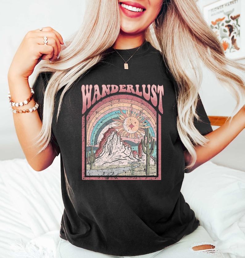 May include: Black t-shirt with a vintage-style graphic of a mountain range, sun, and rainbow. The text "WANDERLUST" is printed in a retro font above the graphic.