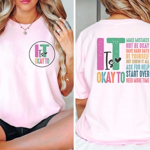 Pode incluir: Camiseta rosa com o texto "It is okay to" em um c&iacute;rculo na frente e o mesmo texto com as palavras "make mistakes, not be okay, have hard days, be yourself, not know it all, ask for help, start over, need more time" nas costas.