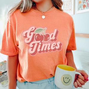 Pu&ograve; includere: T-shirt arancione con la scritta "Good Times" in un font retr&ograve; e una grafica di ciliegie. Una tazza bianca con interno giallo e una faccina sorridente &egrave; tenuta in mano.