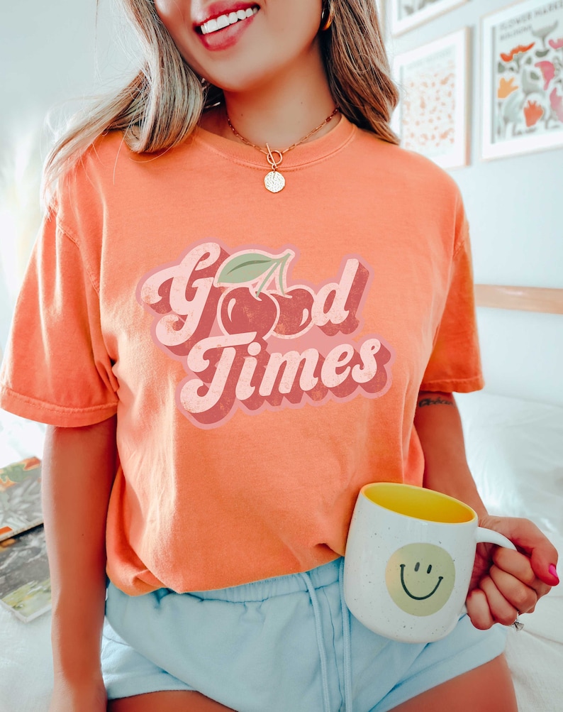 Pu&ograve; includere: T-shirt arancione con la scritta "Good Times" in un font retr&ograve; e una grafica di ciliegie. Una tazza bianca con interno giallo e una faccina sorridente &egrave; tenuta in mano.