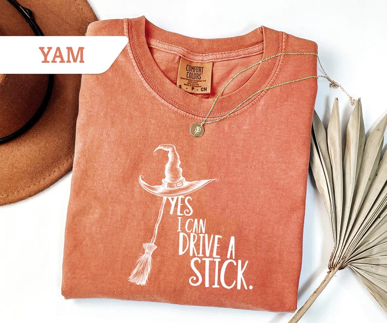 May include: A coral-colored t-shirt with a white graphic of a witch's hat and broom, and the text "Yes I Can Drive A Stick." A gold necklace with a pendant is draped across the shirt. A brown hat is visible in the upper left corner.