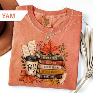 May include: A terracotta-colored t-shirt featuring a graphic print of a stack of books labeled Pumpkin Spice, Crisp Air, Autumn Stories, Sweater Weather, and Cozy Days, alongside a coffee cup with the text Fall and a large maple leaf.