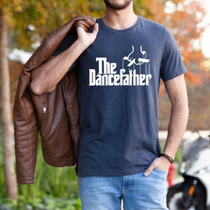 Pode incluir: Uma camiseta azul escura com texto branco que diz "The Dancefather" e um gr&aacute;fico de uma m&atilde;o segurando cordas presas a um fantoche. A pessoa que est&aacute; usando a camiseta est&aacute; segurando uma jaqueta de couro marrom.