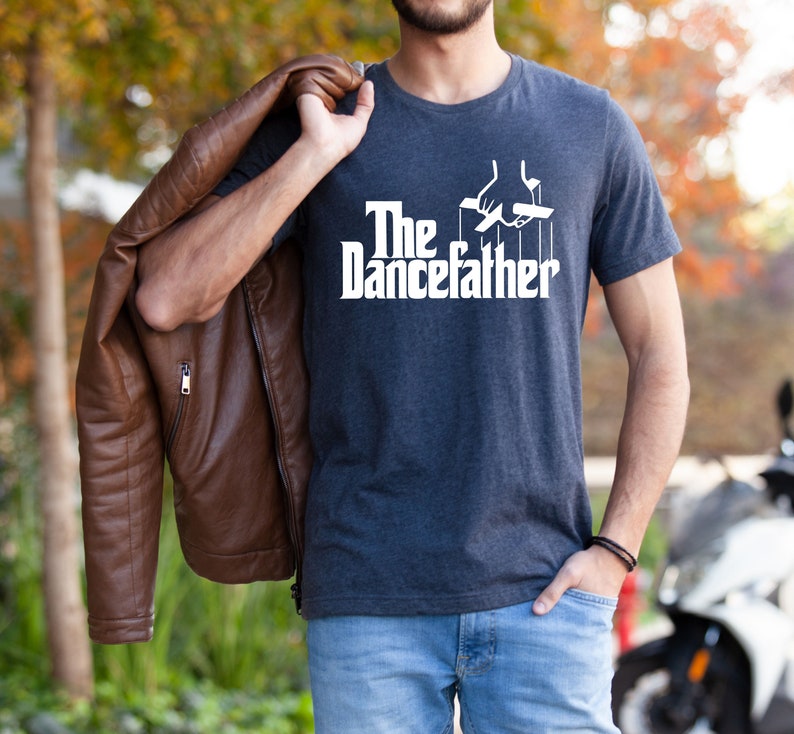 Pode incluir: Uma camiseta azul escura com texto branco que diz "The Dancefather" e um gr&aacute;fico de uma m&atilde;o segurando cordas presas a um fantoche. A pessoa que est&aacute; usando a camiseta est&aacute; segurando uma jaqueta de couro marrom.