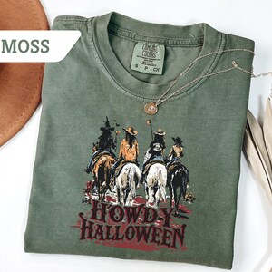 May include: A moss green t-shirt featuring a graphic of four riders on horseback with the text "HOWDY HALLOWEEN" in red. A gold necklace and a brown hat are also visible. The shirt is made of a soft, comfortable material.