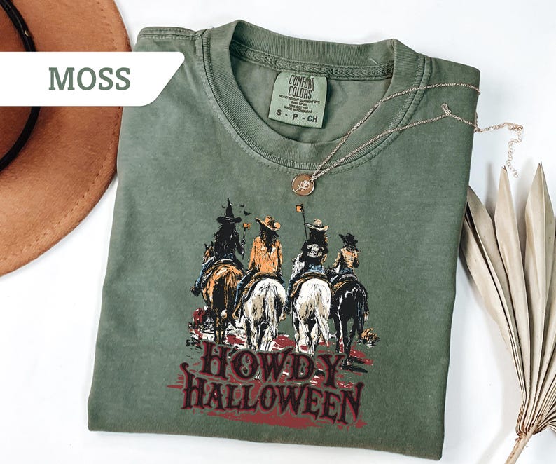May include: A moss green t-shirt featuring a graphic of four riders on horseback with the text "HOWDY HALLOWEEN" in red. A gold necklace and a brown hat are also visible. The shirt is made of a soft, comfortable material.