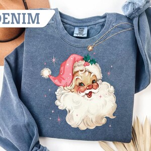 May include: Denim blue sweatshirt featuring a vintage Santa Claus graphic with a pink hat and white beard. The design includes pink stars. The sweatshirt has a crew neck and long sleeves. The word "DENIM" is in the upper left corner.