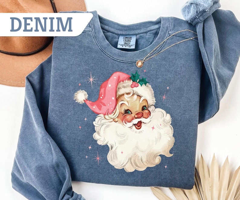 May include: Denim blue sweatshirt featuring a vintage Santa Claus graphic with a pink hat and white beard. The design includes pink stars. The sweatshirt has a crew neck and long sleeves. The word "DENIM" is in the upper left corner.