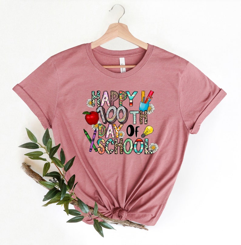 Puede incluir: Una camiseta color malva con el texto "Happy 100th Day of School" en letras de colores decoradas con &uacute;tiles escolares y flores.