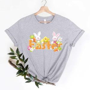 May include: A gray t-shirt with a graphic of two bunnies and a chick holding carrots and flowers. The word "Easter" is spelled out in orange letters with pink accents.