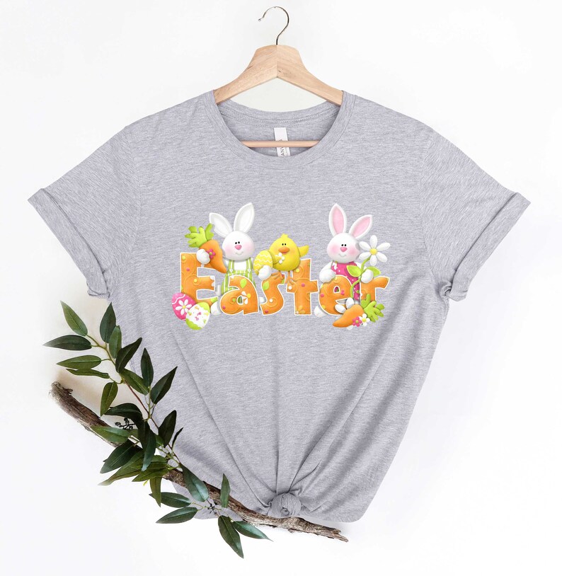 May include: A gray t-shirt with a graphic of two bunnies and a chick holding carrots and flowers. The word "Easter" is spelled out in orange letters with pink accents.